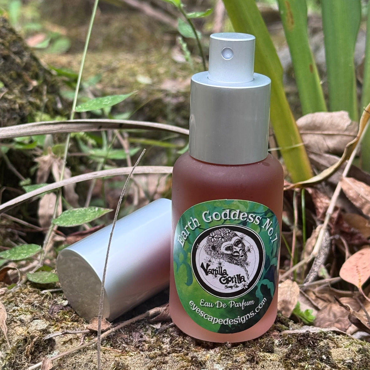 A small glass perfume bottle labeled 'Earth Goddess No.05 Vanilla Gorilla' with a silver spray top, photographed outdoors on a forest floor with dry leaves and green plants around it.