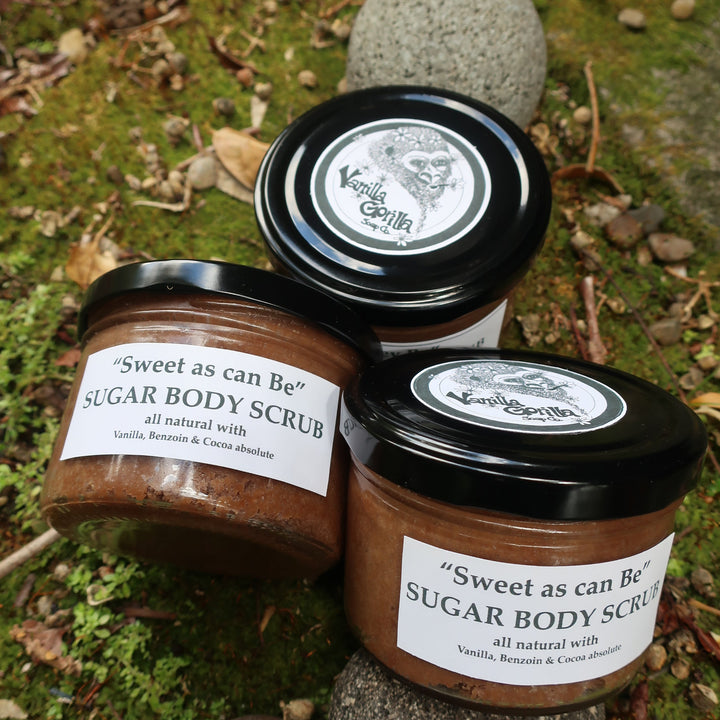 Three jars of 'Sweet as can be' sugar body scrub on a natural stone surface with greenery.