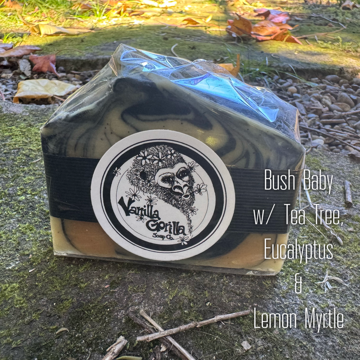 A rectangular bar of soap wrapped in clear cellophane and tied with a black elastic band, placed outdoors on a mossy stone surface. The soap has a marbled black, gray, and tan pattern, with a round black-and-white label reading 'Vanilla Gorilla Soap Co.