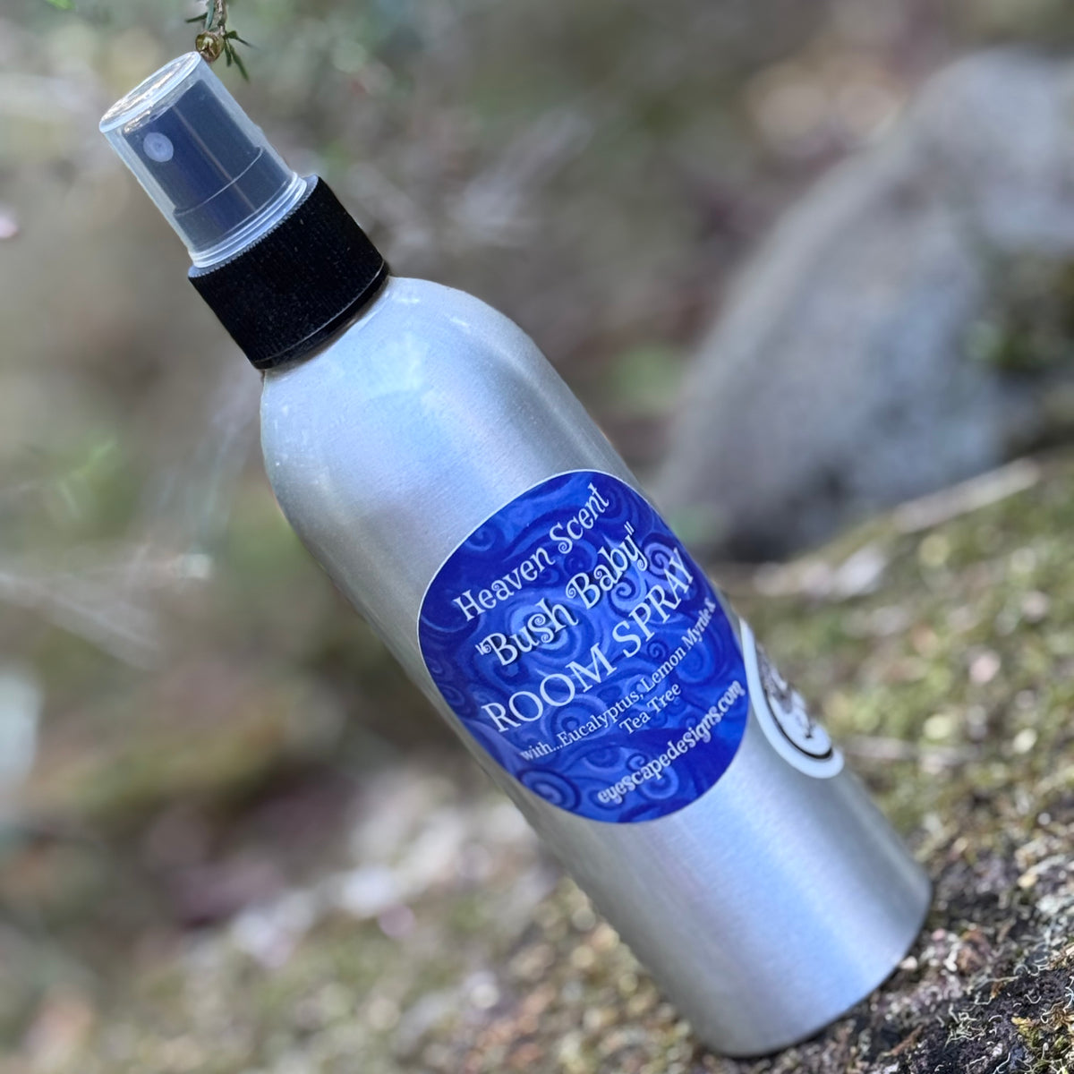 A silver aluminum room spray bottle with a black spray cap, resting on a mossy rock outdoors. The bottle features a prominent blue circular label that reads 'Heaven Scent "Bush Baby" ROOM SPRAY'.