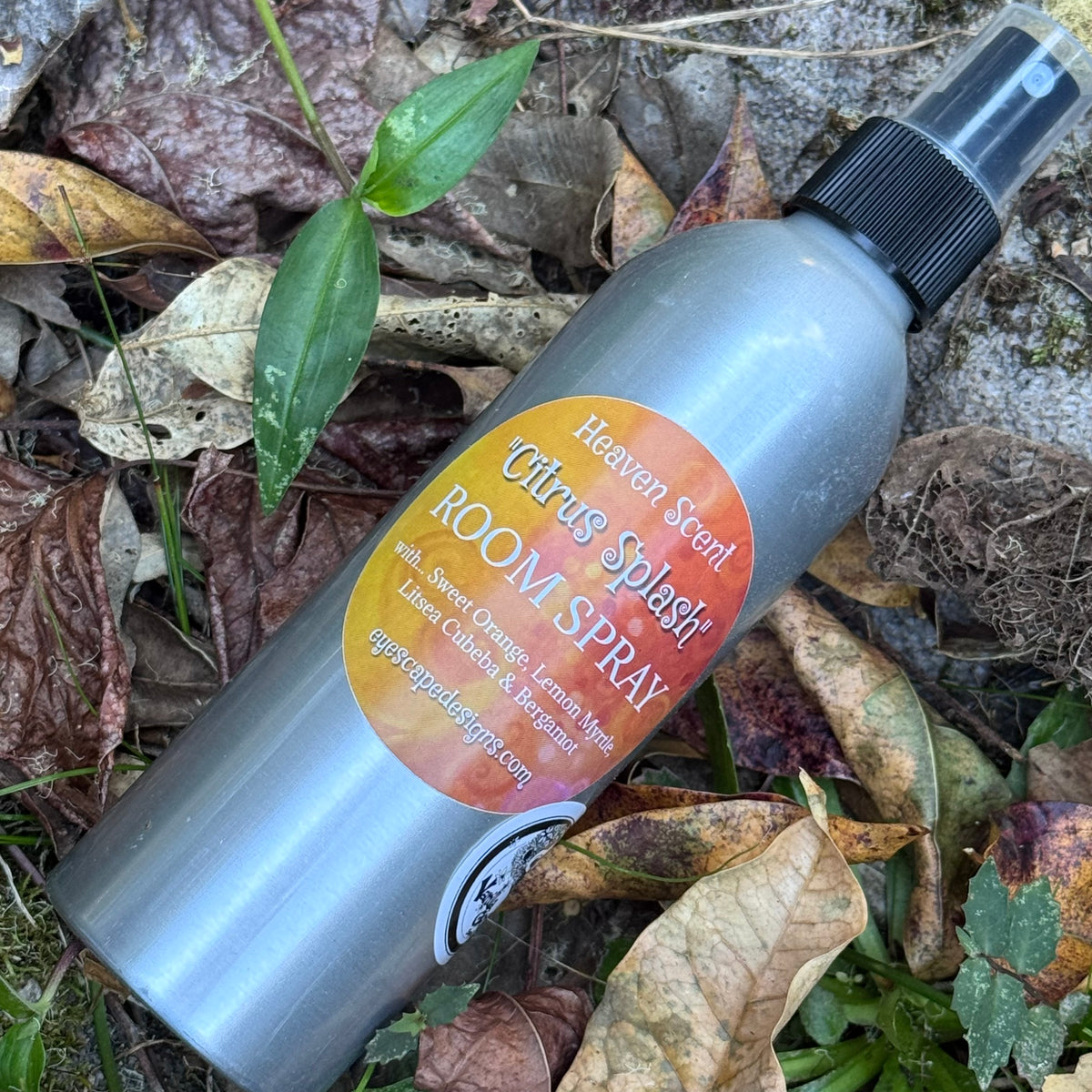 A silver room spray bottle labeled 'Heaven Scent Citrus Splash Room Spray' lying on a bed of dry brown leaves and green grass outdoors.
