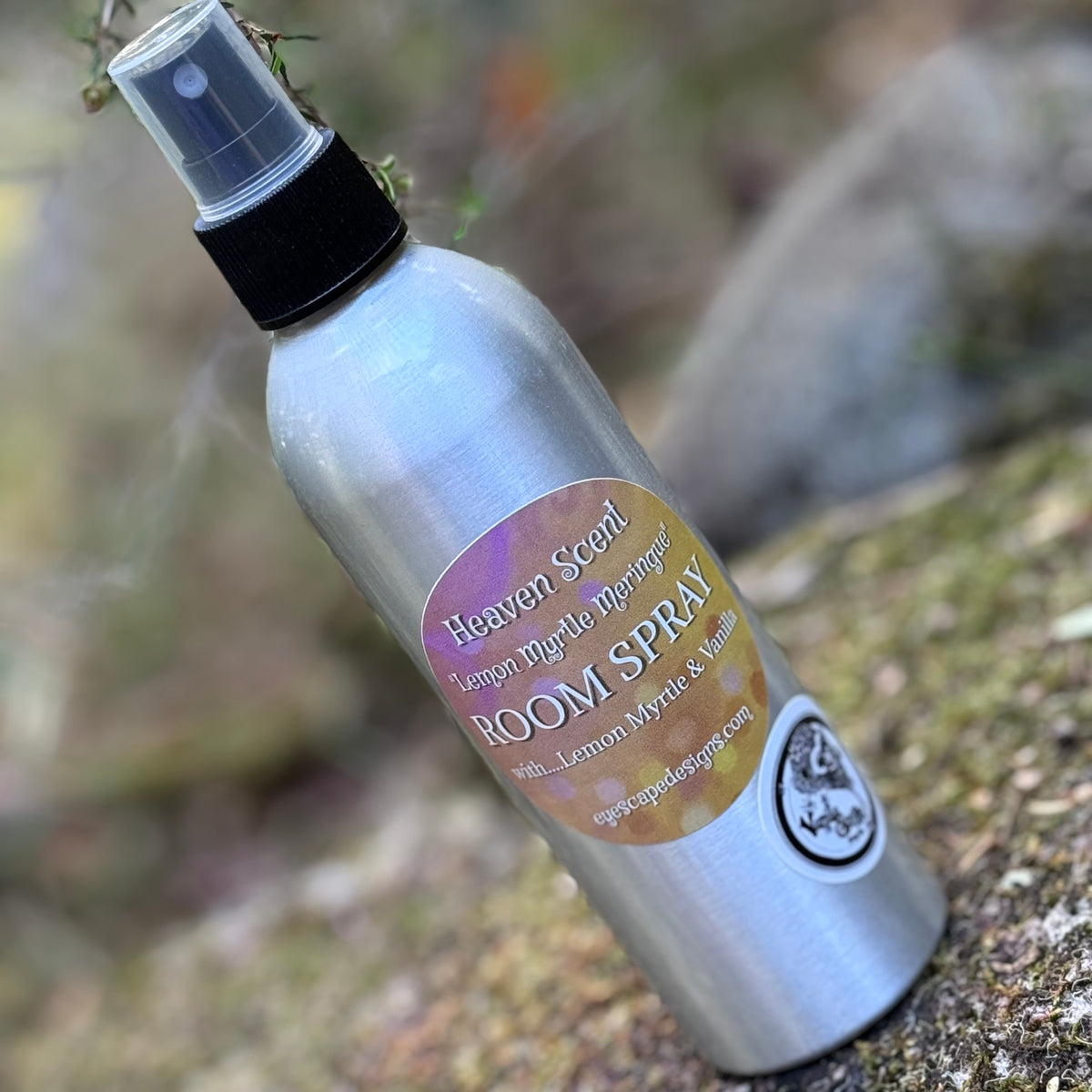 A silver aluminum room spray bottle labeled 'Heaven's Scent Lemon Myrtle meringue' and 'ROOM SPRAY' with a black spray top, photographed outdoors on a mossy rock with a blurred natural background.