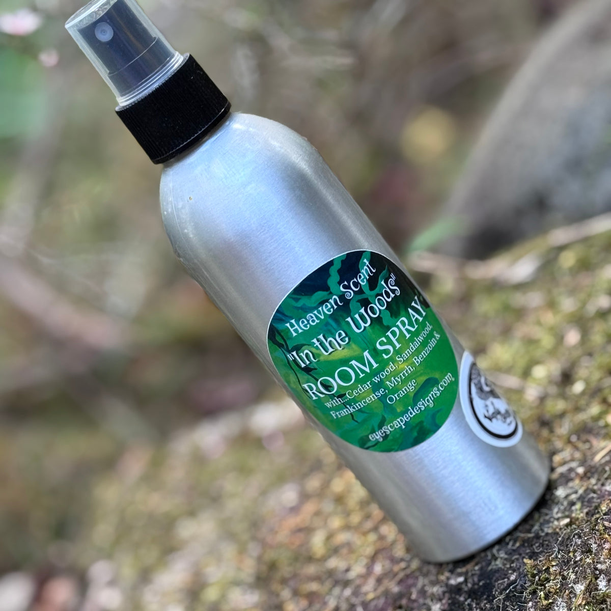 A silver aluminum room spray bottle labeled 'Heaven Scent - In the Woods' sandalwood Cedarwood frankincense bergamot & myrrh with a green and black label, resting on a mossy log outdoors.