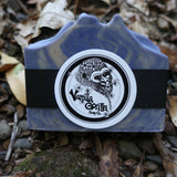 Blue soap bar and black product packaging on a natural background with leaves and stones.