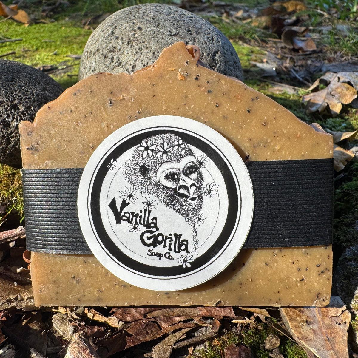 A rectangular bar of soap in a warm tan-brown color with dark flecks, wrapped with a black elastic band and a round white sticker that reads 'Vanilla Gorilla Soap Co.' with a gorilla illustration.