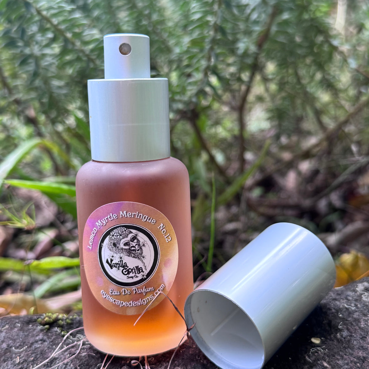 A bottle of Lemon Meringue No.10 Eau De Parfum, featuring a peach-colored translucent bottle, a silver spray pump, and a detached cap lying on a stone surface with a blurred forest background.