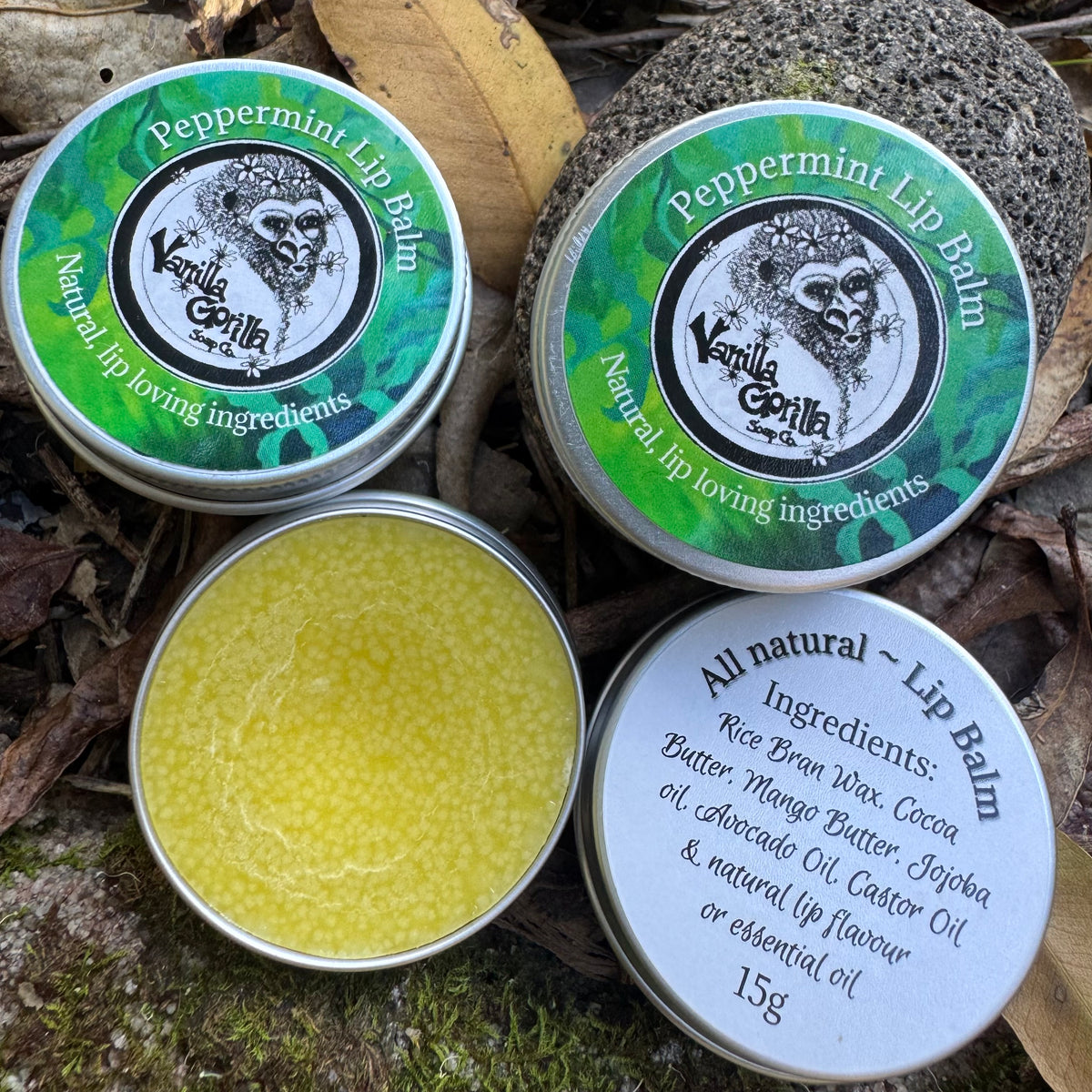 Four round metal tins of Vanilla Gorilla Peppermint Lip Balm arranged on a natural forest floor with moss, leaves, and bark. One tin is open, showing the pale yellow balm inside, while the others show the lids with product labels and an ingredients list.