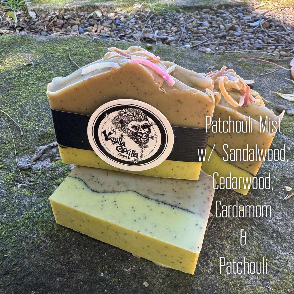 Three artisanal soap bars stacked outdoors on a mossy stone surface. The top bar is tan with pink and orange botanical flecks and a black band; the middle bar is pale yellow with dark speckles; the bottom bar is pale yellow with gray marbling.