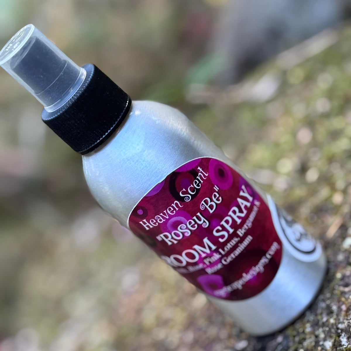 A silver room spray bottle with a black spray cap and a magenta label reading 'Heaven Scent Rosey Be ROOM SPRAY', photographed outdoors on a blurred natural background.