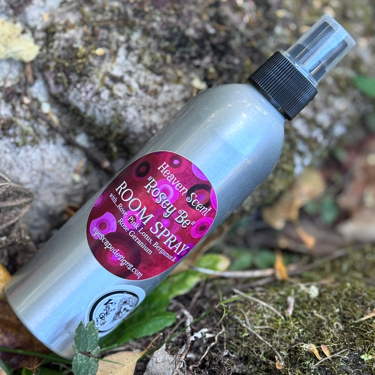 A silver room spray bottle labeled 'Heaven Scent' and 'Rosey be' with rose geranium scent, placed outdoors on mossy ground with leaves.