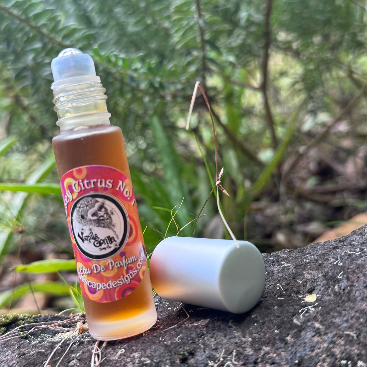 A small roll-on bottle of citrus note fragrance oil with its cap off, placed on a tree trunk outdoors with green foliage in the background. The label reads 'Citrus Note' and 'Eau De Parfum'.