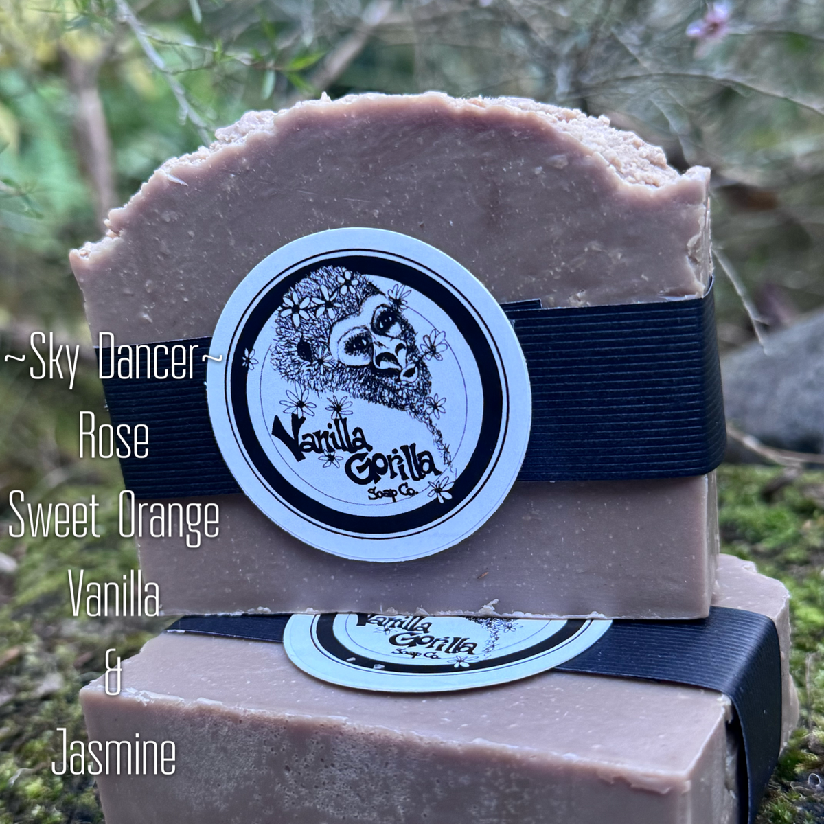 Two handmade soap bars stacked on top of each other outdoors, wrapped with black ribbons and a round white label reading Vanilla Gorilla Soap Co. The top bar is labeled “~Sky Dancer” and the visible scent notes on the left read “Rose Sweet Orange Vanilla & Jasmine.