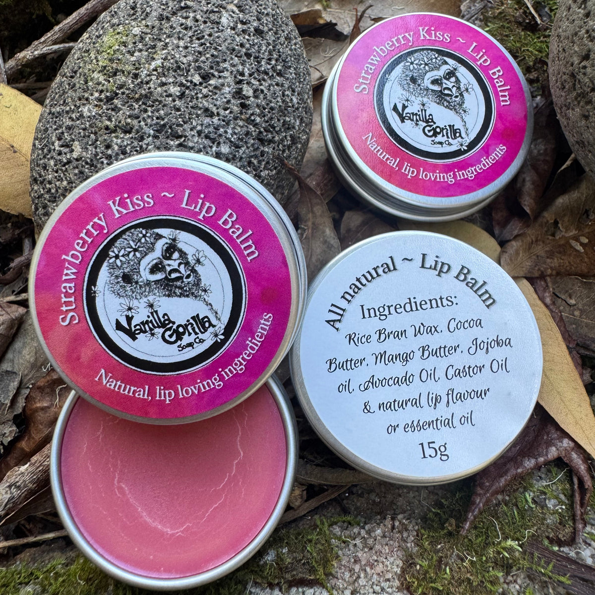 Three small round metal tins of lip balm arranged on moss and dry leaves outdoors. One tin is open, showing pink balm inside, and two tins are closed with bright pink labels.