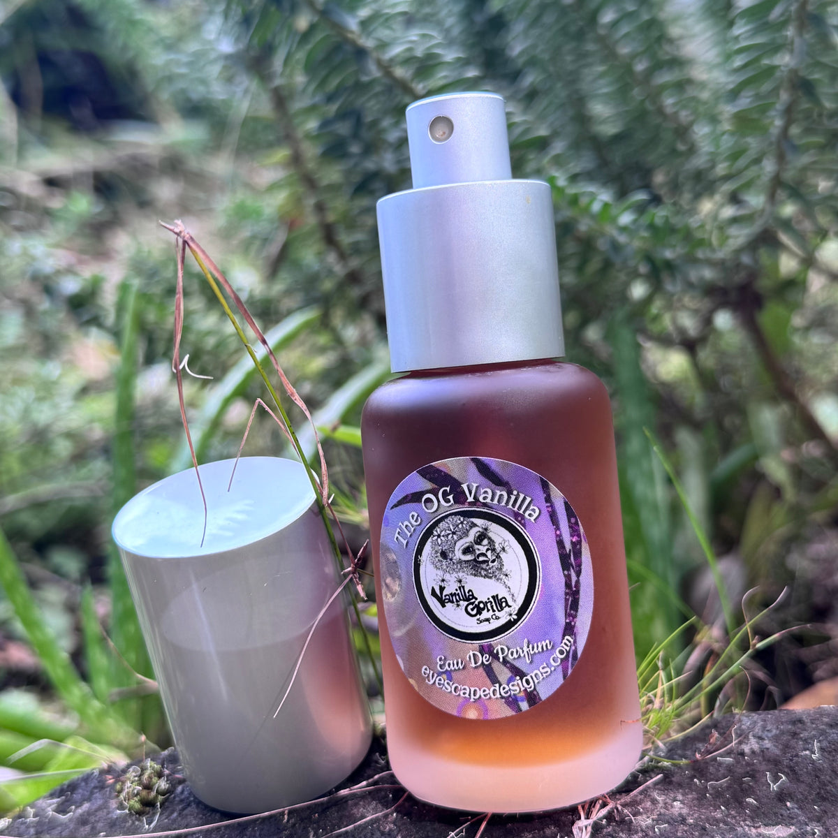 A bottle of OG Vanilla Eau De Parfum by The Earth & Vine next to its silver cap, set outdoors on a log with green foliage in the background.