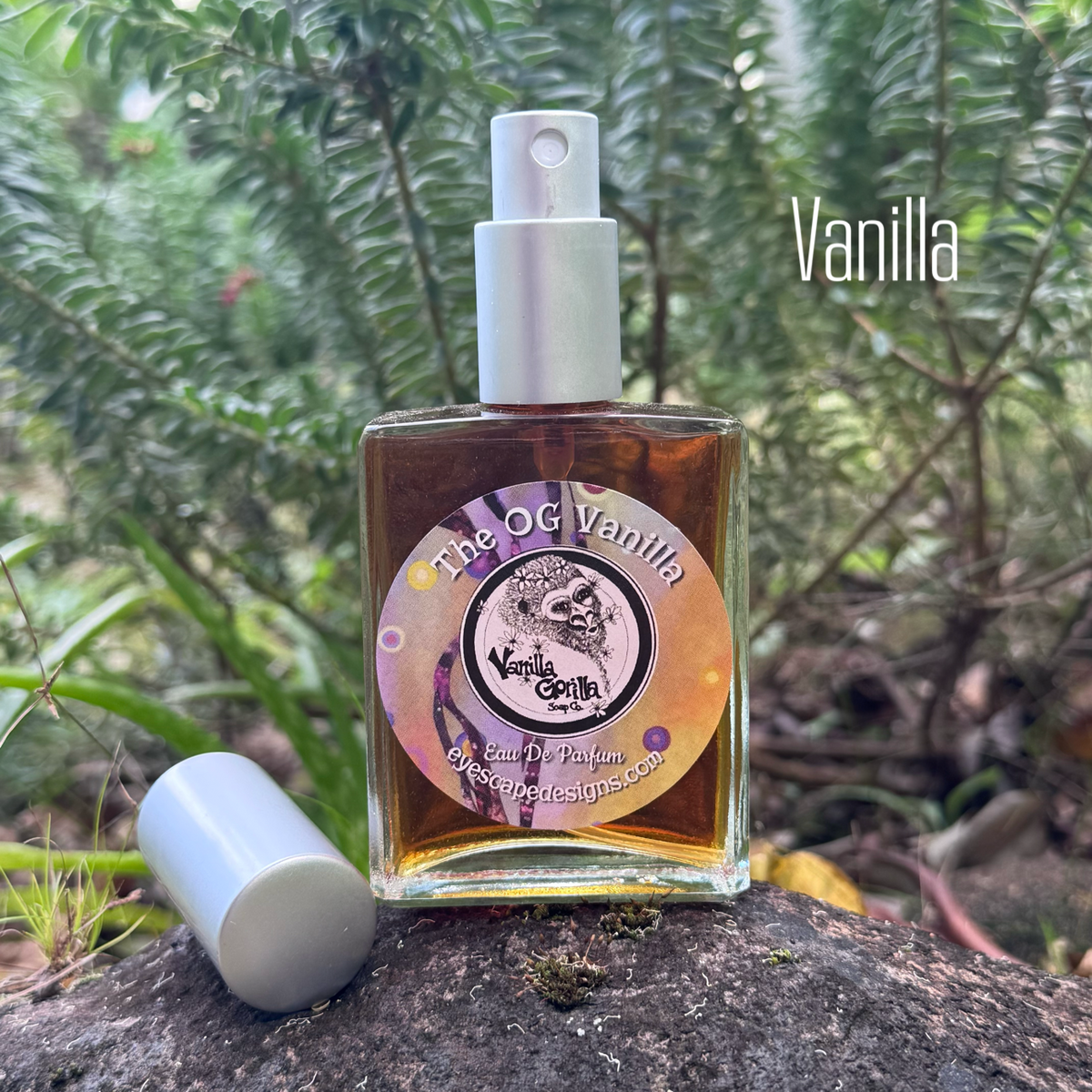 A bottle of 'The OG Vanilla' essential oil fragrance by 'vanilla gorilla' sits on a dark rock with its cap placed beside it. The background is a lush, out-of-focus green forest.