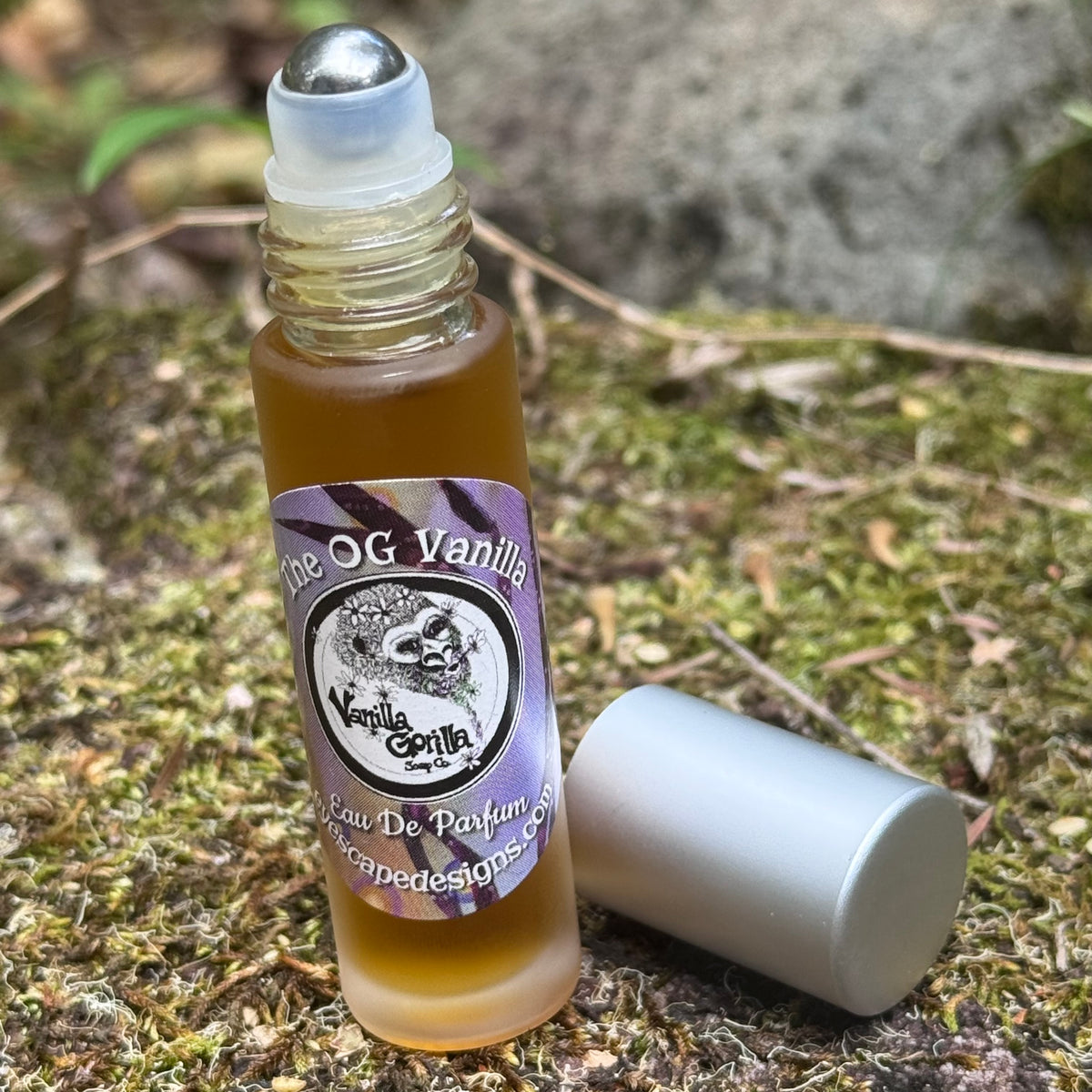 A small glass roller bottle filled with amber liquid stands on mossy ground outdoors, with its silver cap placed beside it. The label reads 'The OG Vanilla' and 'Vanilla Gorilla' Eau De Parfum.