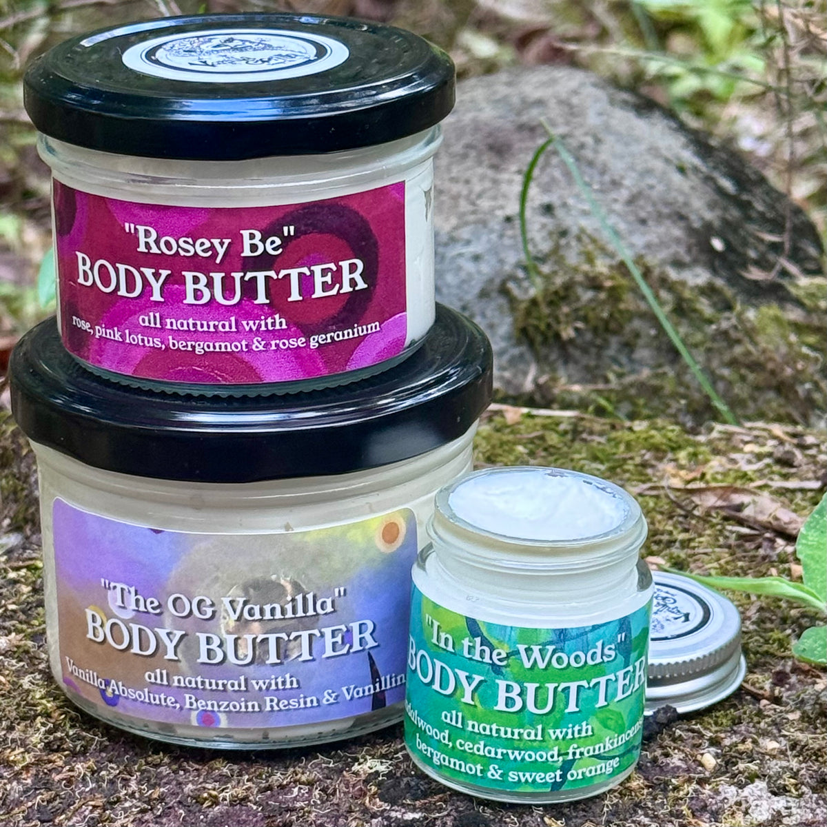 Three jars of body butter displayed outdoors on a mossy rock surface. The stack includes a small jar labeled "Rosey Be" at the top, a larger jar labeled "The OG Vanilla" in the middle, and an open jar labeled "In the Woods" at the front right.