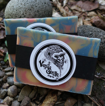 Colorful orange swirl soap bars with a black strap and vanilla gorilla label on a textured surface with leaves and stones.