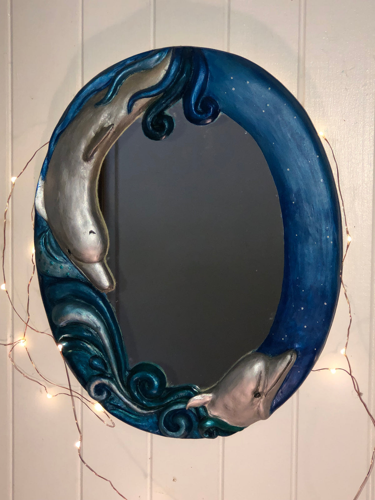 Oval mirror with 2dolphins sculpted around in blue sea on a cream wall with fairy lights